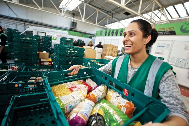A woman with her hair pulled back in a bun is smiling and holding up a green container of food at a food bank.
